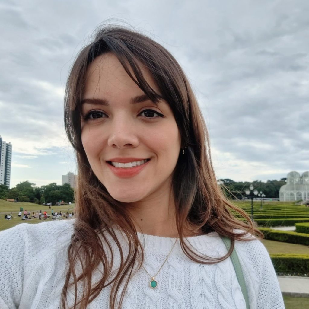 Woman smiling, wearing a white sweater and a necklace, in the foreground. In the background, there is a large garden with geometric hedges and a glass structure, with people walking around under a cloudy sky.