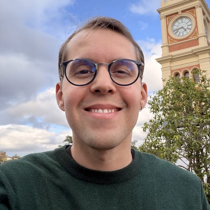 Henrique Aveiro smiling, wearing glasses and a green sweater in the foreground. In the background, there is a tall clock tower with beige and red details, surrounded by trees and a blue sky with clouds.